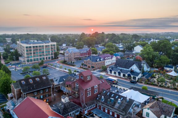 Aerial view of a small town at sunrise with mixed architectural styles, including a multi-story modern building, traditional houses, and a street grid layout, with the sun rising in the hazy sky in the background.