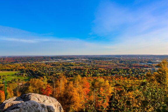 A scenic autumnal vista overlooking a vibrant landscape with colorful fall foliage, a clear sky, and a distant urban area.