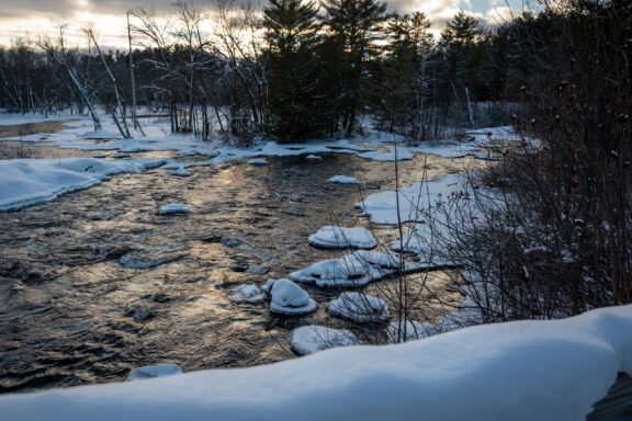 A snow-covered river landscape during winter with patches of ice on the water, surrounded by bare trees and evergreen forest, under a partly cloudy sky at dusk.