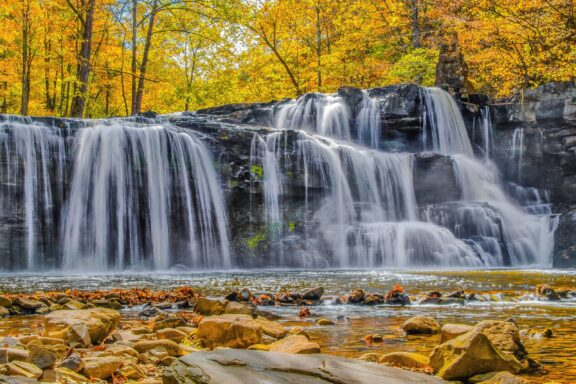 A scenic autumn waterfall with golden foliage in the background and rocky terrain in the foreground.