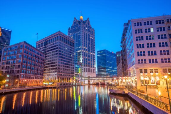 Twilight view of an urban cityscape with illuminated office buildings reflected in a calm river, featuring a prominent tower with a pitched roof and flag.