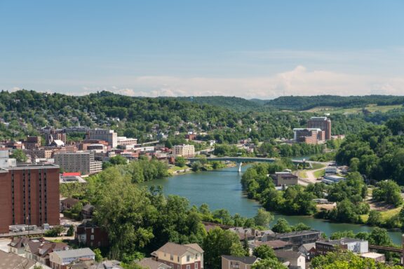 A panoramic view of a town with buildings spread across rolling hills, a river meandering through, bridges crossing over the water, and lush greenery under a clear blue sky.