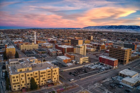 Aerial view of a cityscape during sunset with illuminated buildings, long shadows, and a mountain range in the background.