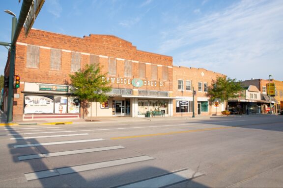 A sunny view of a quiet main street with a pedestrian crosswalk, showcasing a row of old brick buildings with various storefronts including a drug store.