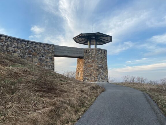 A pathway leading towards an observation tower with stone pillars and an overhanging roof against a cloudy sky background.