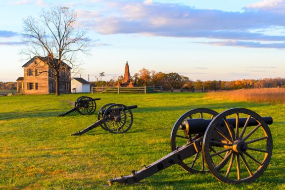 A historic battlefield scene at sunset with antique cannons in the foreground, a wooden farmhouse, and a distant memorial obelisk against a backdrop of a vibrant sky and scattered clouds.