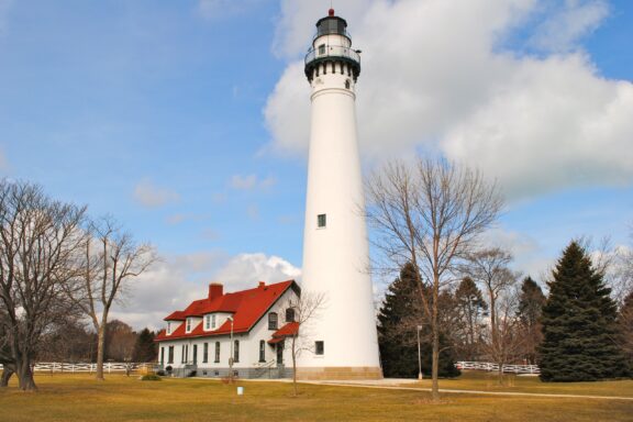 A white lighthouse with a green lantern room stands tall beside a house with a red roof on a clear day with scattered clouds. Trees and a white fence surround the property on a grassy field.