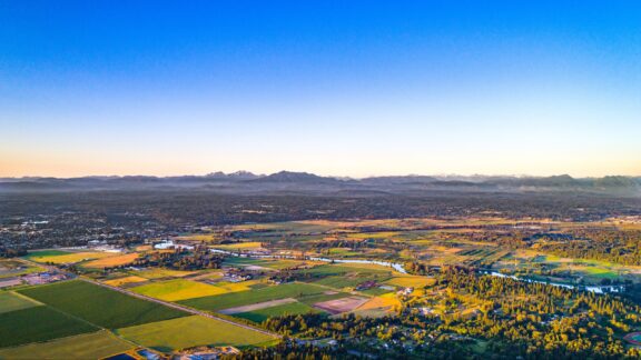 Aerial view of a sprawling landscape at sunset with patchwork fields in the foreground and a mountain range in the distance under a clear blue sky.