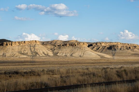 A vast, arid landscape with a railroad track in the foreground, expansive grassland leading up to rugged desert hills under a blue sky with scattered clouds, and an electricity pylon standing in the middle distance.