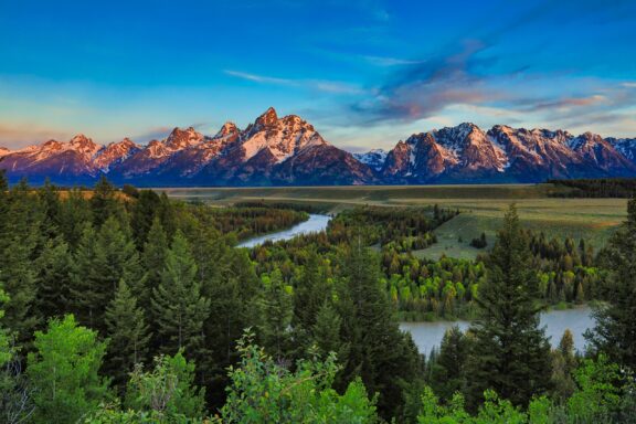 A stunning view of the Teton Range at sunrise with a winding river in the foreground surrounded by lush greenery.