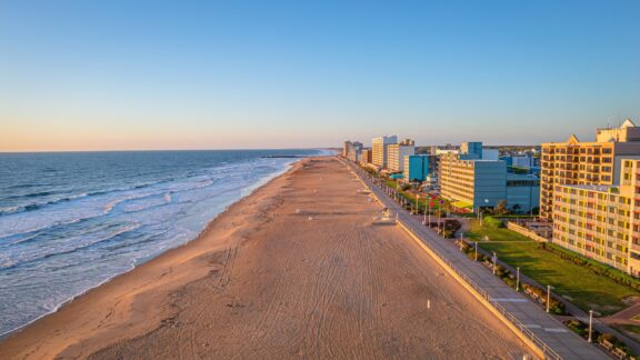 Aerial view of a sunlit beach alongside a road with a row of hotels, under a clear blue sky during early evening.