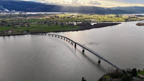 Aerial view of a curved bridge spanning a wide river with patches of sunlight illuminating the surrounding landscape and hills in the distance.