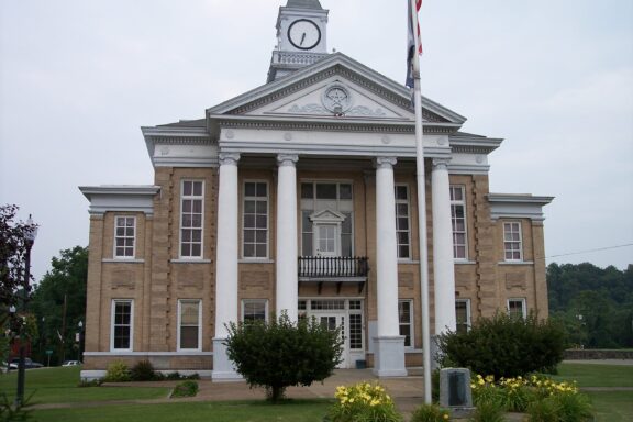 A neoclassical courthouse with a prominent clock tower, tall white columns, and an American flag in front. Landscaped with grass and flower beds under a cloudy sky.