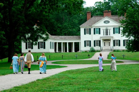 A group of people in historical clothing walking towards a large two-story white colonial house with green shutters and a covered porch, surrounded by lush green trees and grass.