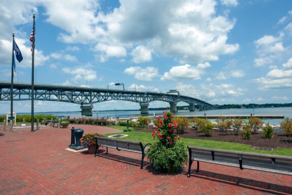 A waterfront park with benches, flowerbeds, and flags in the foreground with a view of a long steel truss bridge extending over a wide river under a partially cloudy sky.