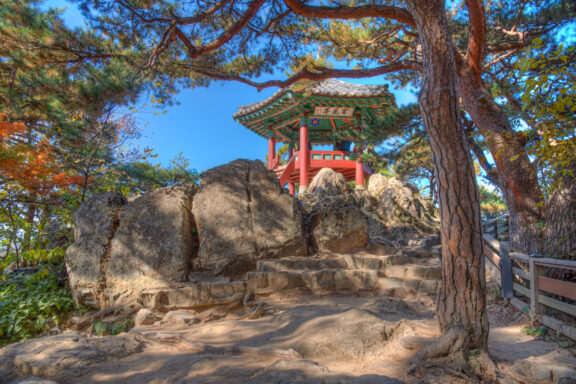 A traditional Korean pavilion nestled on a rocky outcrop surrounded by pine trees under a clear blue sky.