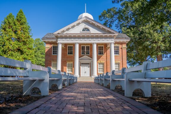 A brick pathway leading to a colonial-style building with white columns, a central pediment, and a cupola, flanked by white benches and green trees under a clear blue sky.