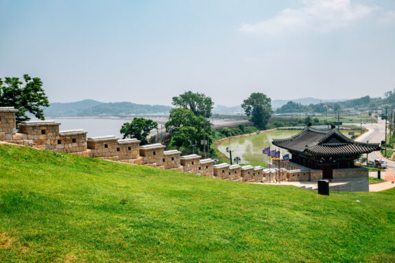 A traditional Korean pavilion next to a stone wall on a grassy hillside, with a river in the background and clear skies overhead.