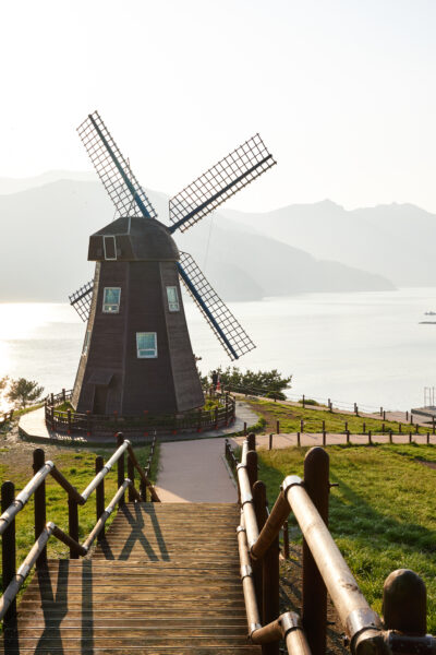 A traditional wooden windmill on a hill overlooking a lake with mountains in the background during sunset.