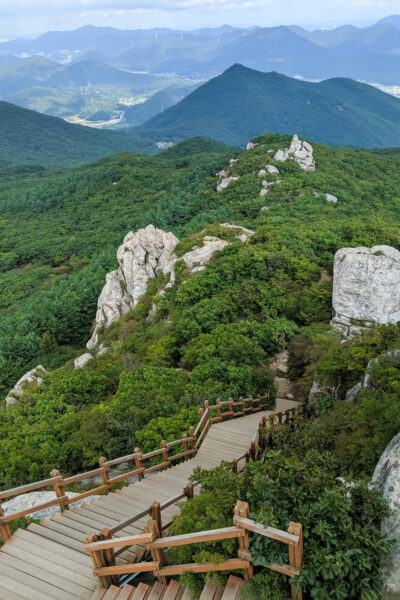 A wooden staircase winding down a lush mountain slope with large rocks and a panoramic view of distant mountains under a blue sky with clouds.