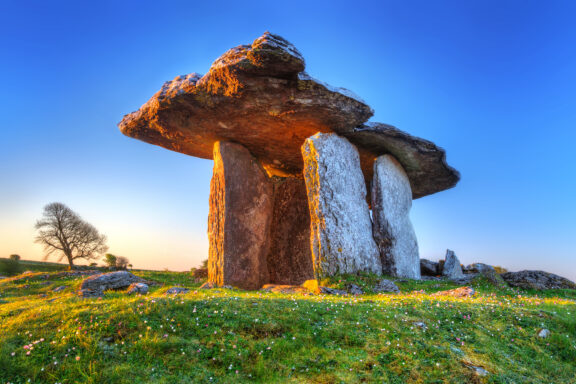 A Neolithic dolmen with a large capstone resting on three upright stones against a clear blue sky during sunset, surrounded by a grassy field with wildflowers and a solitary tree in the background.