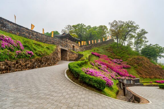 A winding cobblestone pathway leads to an ancient stone gateway adorned with traditional flags, surrounded by lush greenery and vibrant purple azalea bushes.