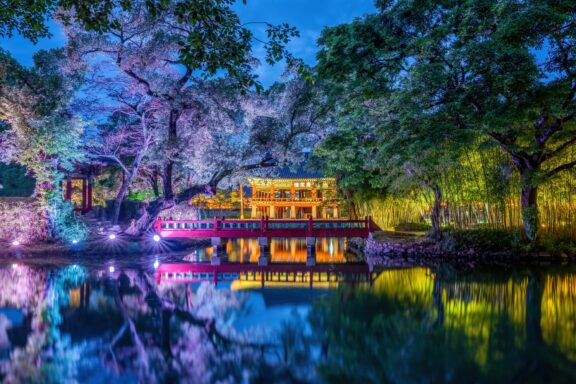 A traditional Asian pavilion illuminated at night, with a reflection in a tranquil pond, surrounded by colorful, lighted trees and bamboo.