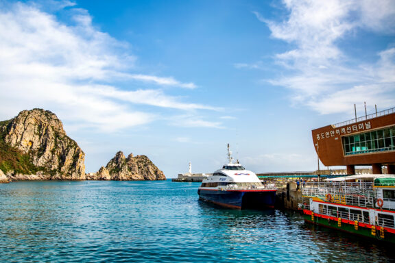 A white and blue high-speed ferry docked at a port with a building displaying Korean text and rugged cliffs in the background under a blue sky with scattered clouds.