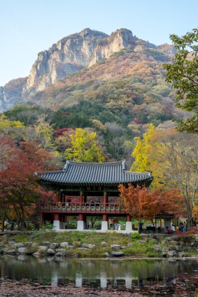 A traditional Korean pavilion by a tranquil pond with autumn-colored trees in the foreground and a rugged mountain in the background.