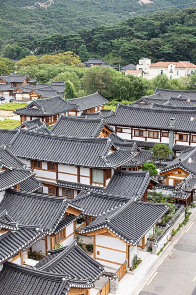 Aerial view of a traditional Korean village with hanok houses featuring tiled roofs and white walls, nestled in a lush green mountainous area.