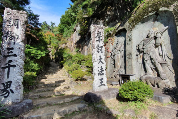 Stone carvings and pillars with engraved text at a serene Japanese temple grounds, surrounded by lush greenery and stairs leading up the hillside.