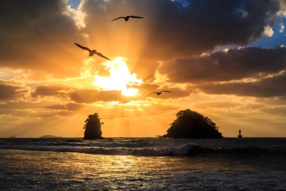 Sunset over the ocean with rays of sunlight piercing through clouds, highlighting rock formations and a lighthouse with seagulls in flight.