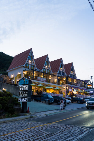 A two-story building with a pitched roof featuring dormer windows illuminated at twilight, with several businesses including a restaurant. There's a cobblestone street in the foreground and signage in Korean. People can be seen walking and several cars are parked in front.