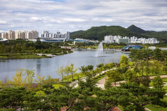 A scenic view of a city park with a large fountain in a lake, surrounded by lush greenery, walking paths, and modern apartment buildings in the background with hills on the horizon.