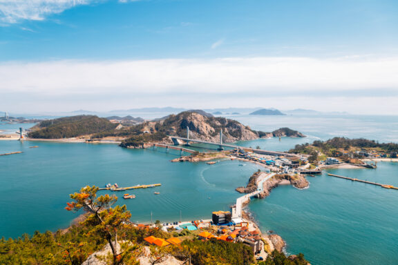 A scenic view of a coastal area with an expansive bridge crossing blue waters, connecting multiple islands and hills under a clear blue sky.