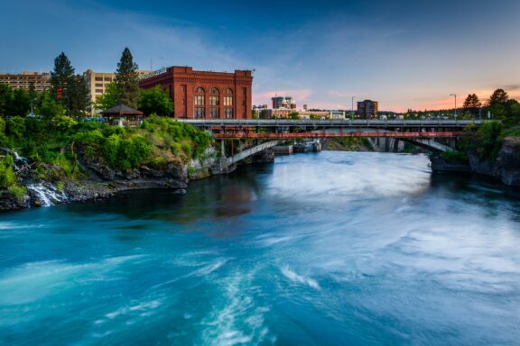 A rushing river flowing under a bridge with a red brick building on the left, amidst an urban backdrop at twilight.