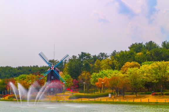 A traditional Dutch windmill beside a lake with multiple fountains, surrounded by colorful autumn trees under a partly cloudy sky.