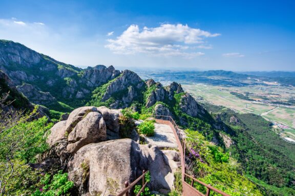 A scenic view from a mountain peak with a metal railing, overlooking a rugged landscape with sharp cliffs, lush greenery, and a vast valley extending into the distance under a bright blue sky with scattered clouds.